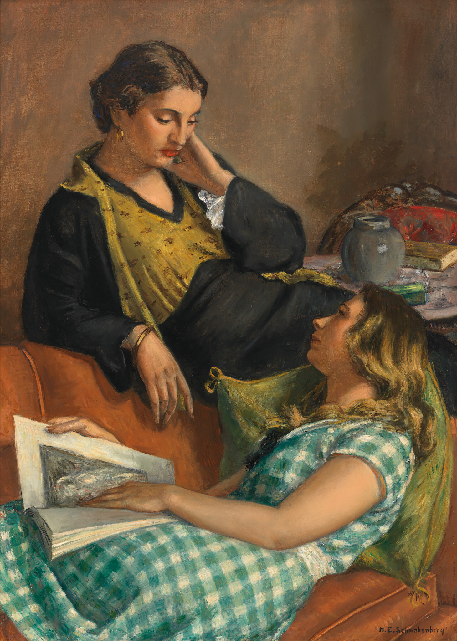 Two women relax on a couch while one reads from a large book and the other watches.