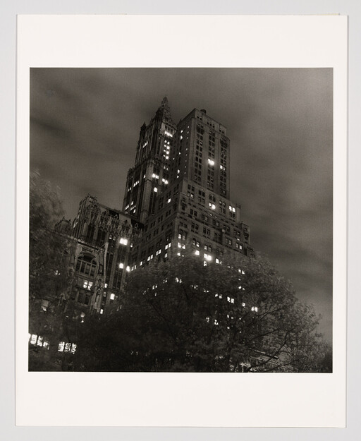 Tall Gothic-style skyscraper rising above trees with many lit windows at night.