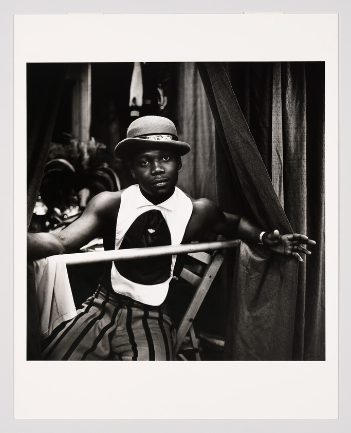 A man in a vintage clown-like costume and bowler hat leans on a railing between curtains.