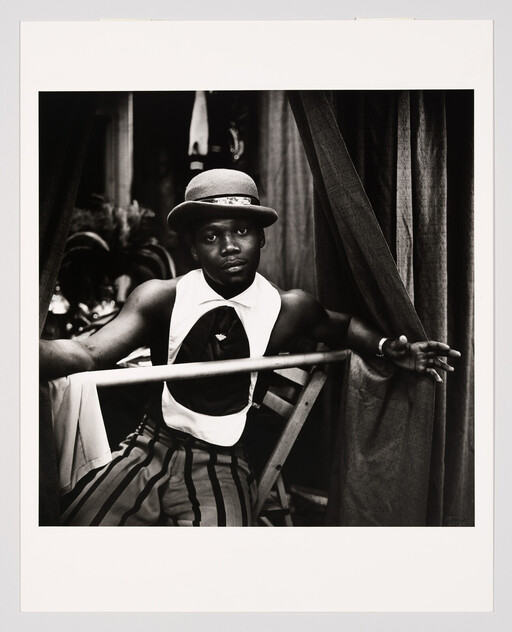 A man in a bowler hat and striped pants leans on a railing by curtains.
