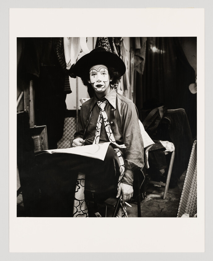 A clown in full makeup and hat sits in a dressing room holding a cigarette and sketchbook.