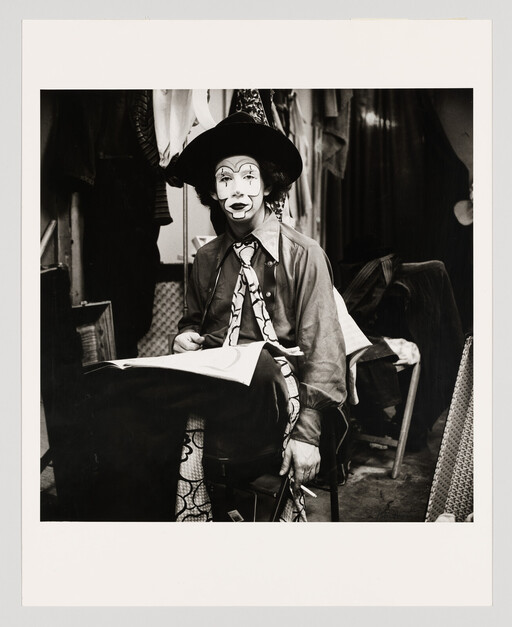 A seated clown with painted face and hat holds a cigarette while sitting with paper on his lap.