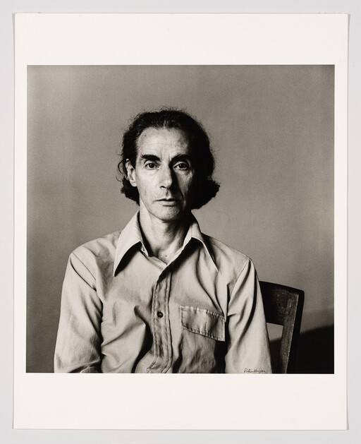A serious middle-aged man sits on a chair wearing a button-up shirt in a black-and-white portrait.