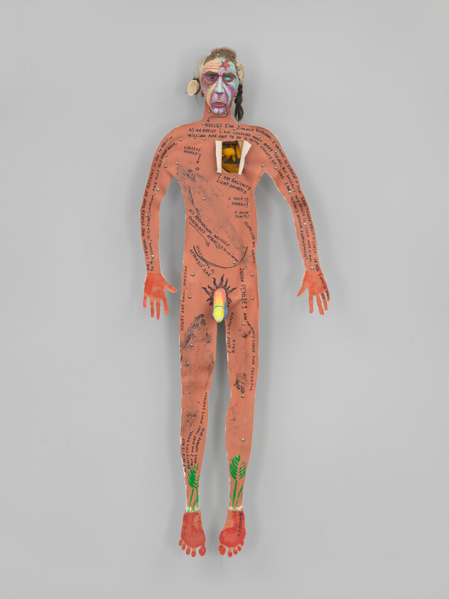 A colorful, humanoid art piece with text and drawings on its body is displayed against a grey background. The figure has a painted face with exaggerated features, and its limbs are spread out. The body is covered with handwritten words and phrases, and there are drawings of green plants on the feet. The art piece has a three-dimensional quality with some elements that appear to be attached or protruding from the surface.