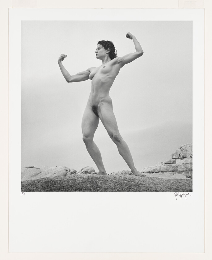 Young nude person standing on a rocky outcrop flexing both arms in a classic strongman pose.