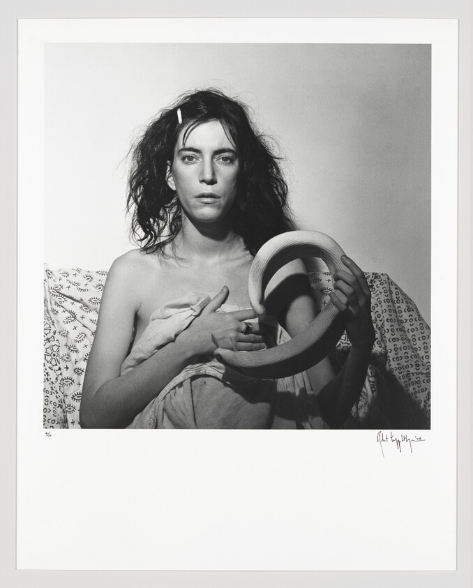 Woman with disheveled hair sits wrapped in cloth while holding a circular wooden ring.