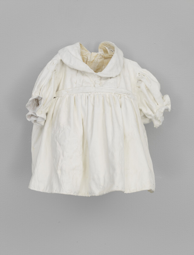 A small vintage white child's dress with puffed sleeves and a sailor collar hangs against a gray wall.