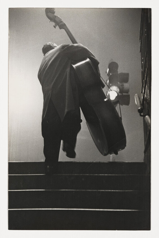 Musician carries a large double bass up a dim stairway toward stage lights.