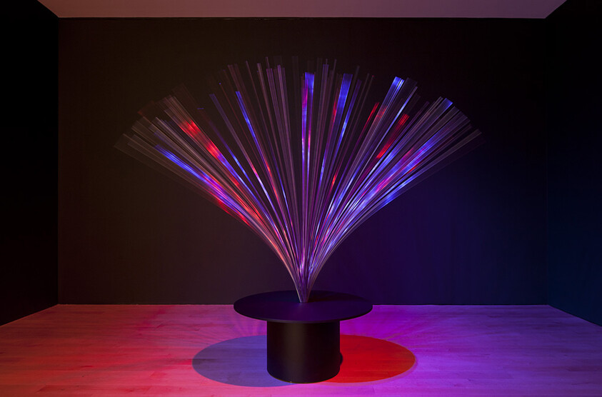 A fiber-optic light sculpture fans colorful blue and red beams from a round black pedestal.