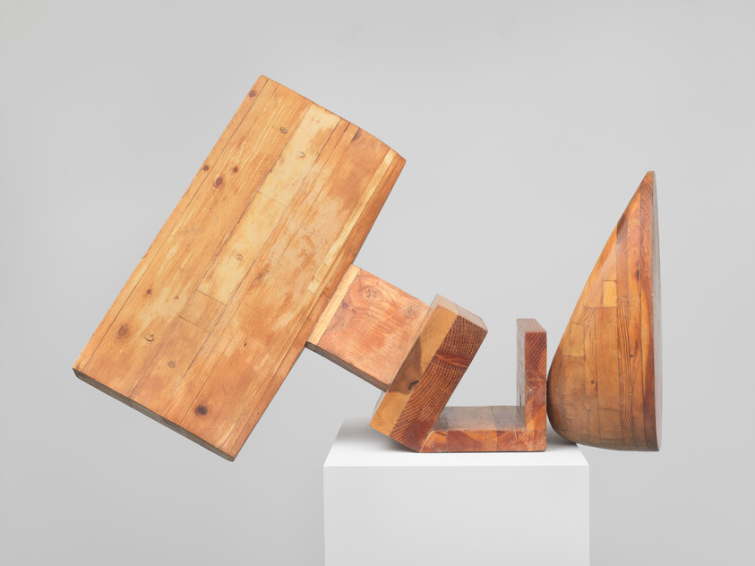 Wooden geometric sculpture with stacked and leaning blocks balanced on a white pedestal.