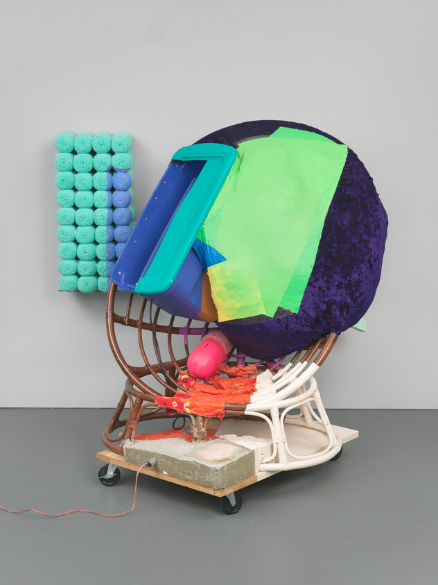 Colorful mixed-media sculpture combines rattan chair, large purple cushion, green fabric, plastic tub on wheels.