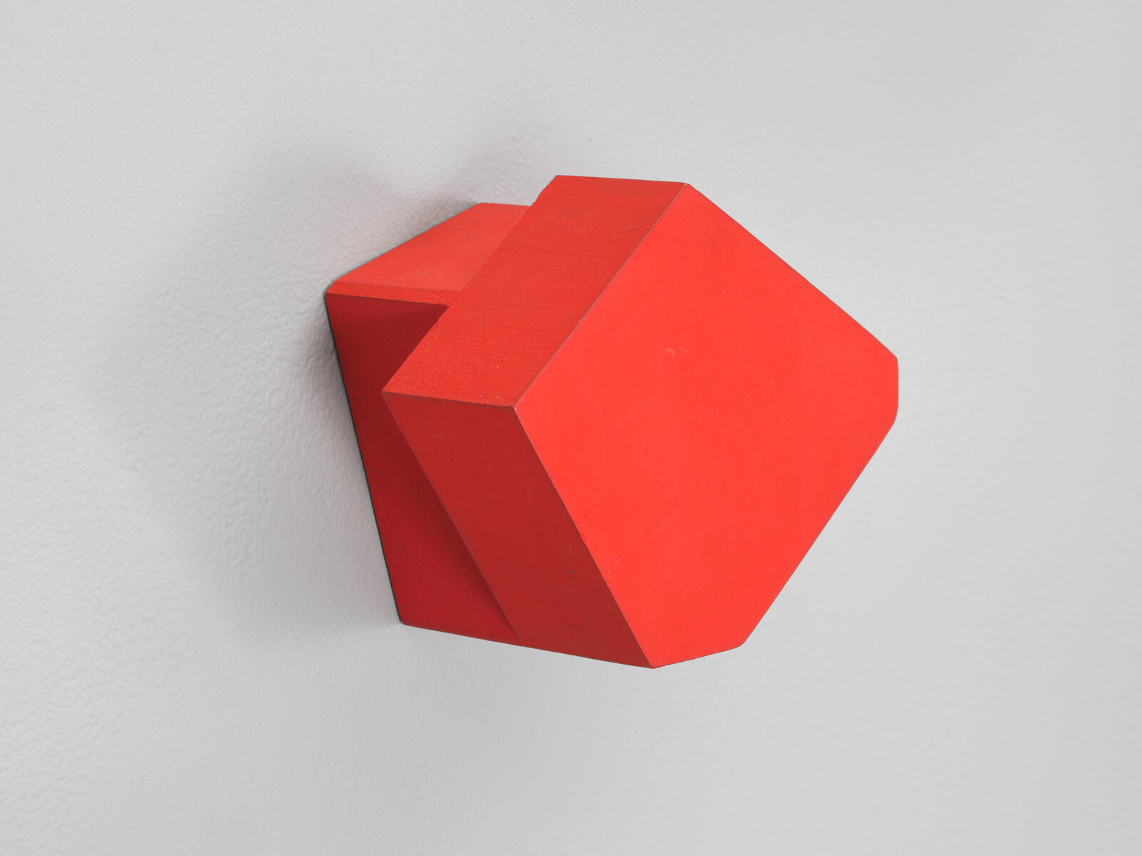 A solid red geometric block with angled sides is attached to a plain light gray wall.