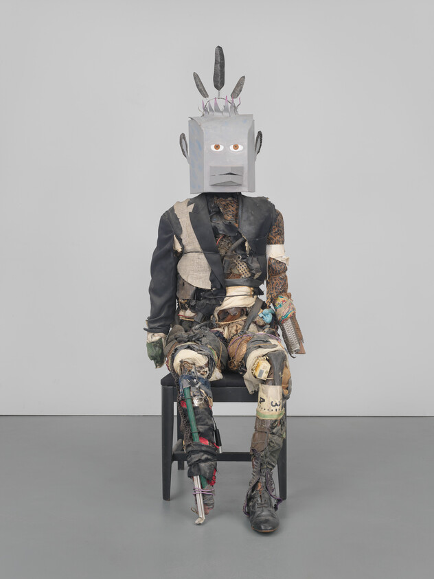 A seated human-like sculpture made from torn clothing and a box-shaped head with feathers.