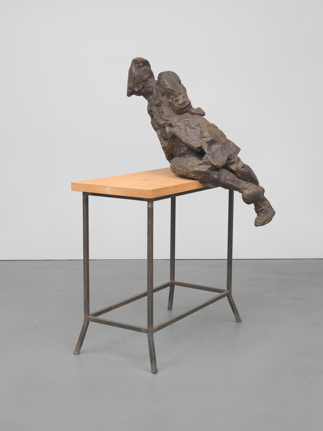 Bronze-like rough human figure leaning backward on the edge of a wooden table.