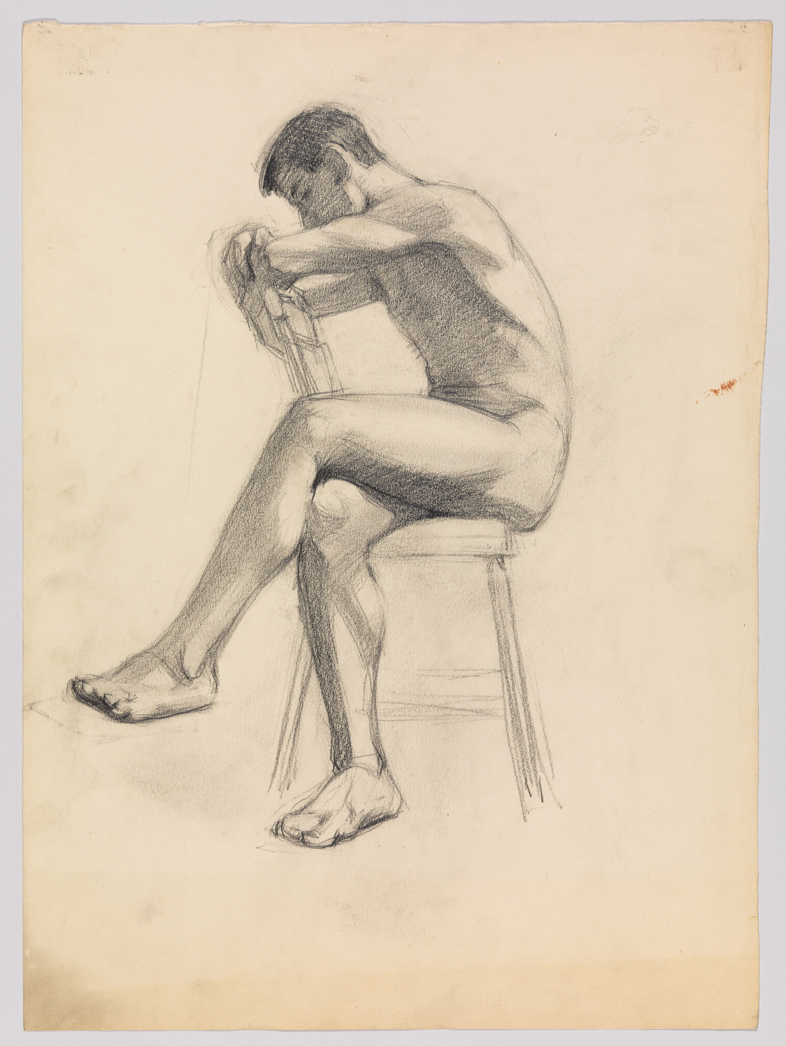 Nude man sitting on a stool leaning forward with arms resting on a chair back.