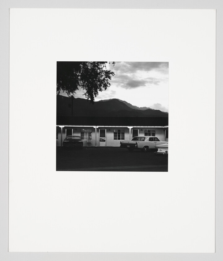 A black-and-white motel with three vintage cars parked in front and mountains behind.