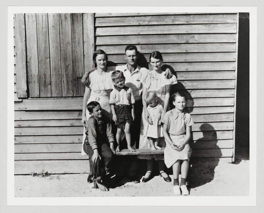 A family of seven poses together on a bench in front of a wooden house.
