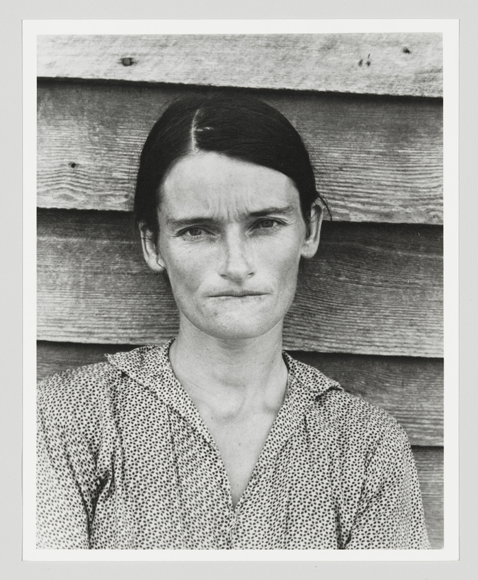 Black and white photo of a solemn woman in a polka dot dress against a wooden backdrop.