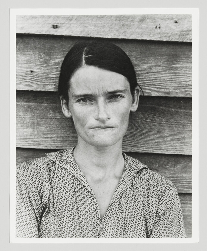 A stern-looking woman with dark hair stands against weathered wooden siding, wearing a patterned blouse.