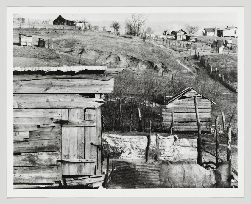 Several weathered wooden shacks and fences sit on a barren hillside with visible soil erosion.