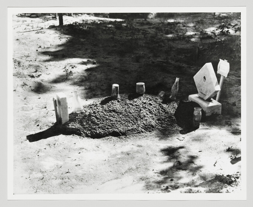 A small dirt grave marked with bottles and makeshift wooden markers under dappled sunlight.