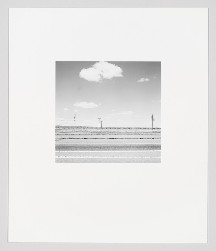 A black-and-white roadside scene with empty highway, distant telephone poles, and a single cloud overhead.