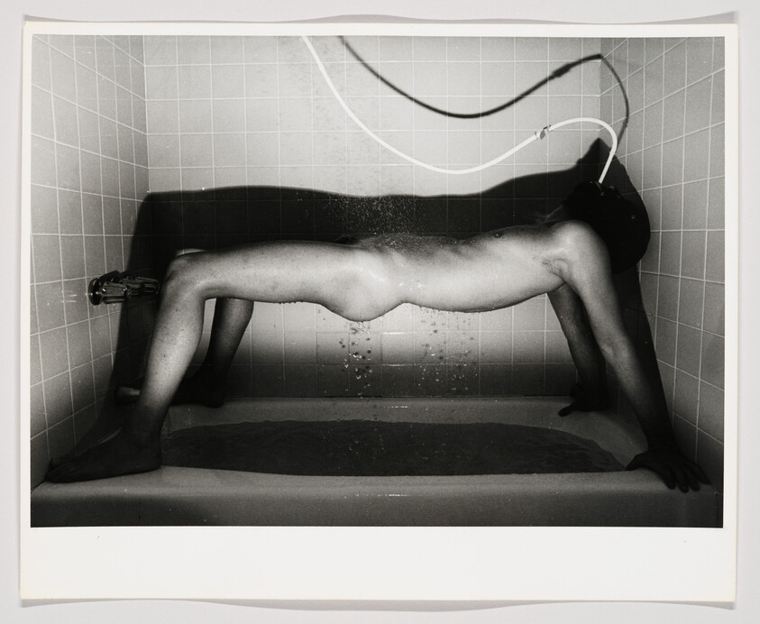 Black and white photo of a person lying in a bathtub with water spraying from a shower head above.
