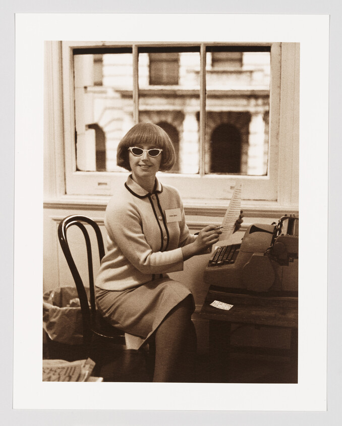 A woman in cat-eye sunglasses sits at a typewriter holding a sheet of paper and smiling.