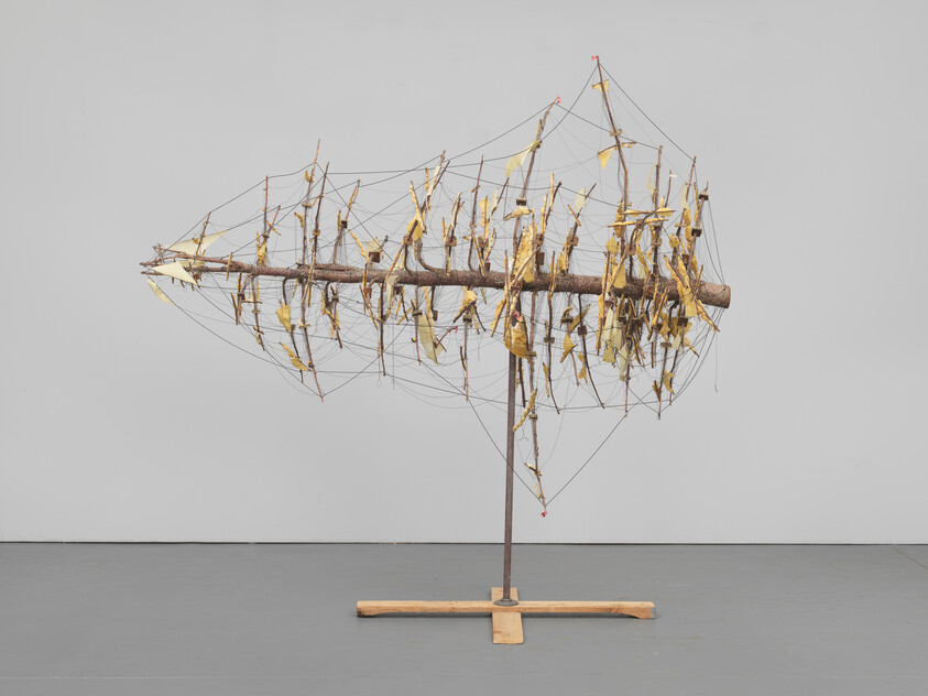 Wooden log sculpture with yellow fabric fragments suspended by many thin wires on a metal stand.