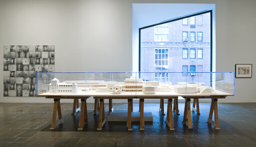 A detailed white architectural model is displayed on a long wooden table under a large window.