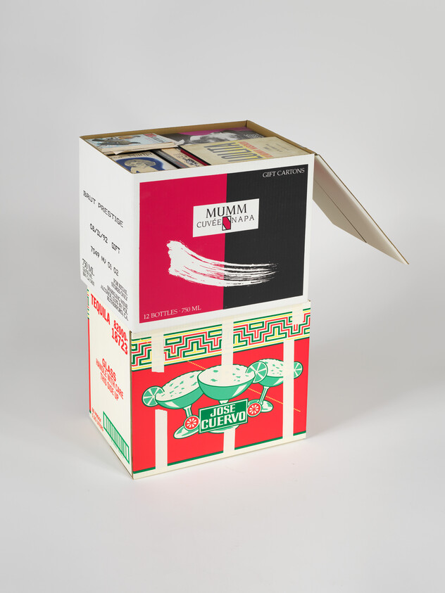 A black and red box of Mumm wine, filled with books, sits on top of a box of a brightly colored red and green Jose Cuervo box