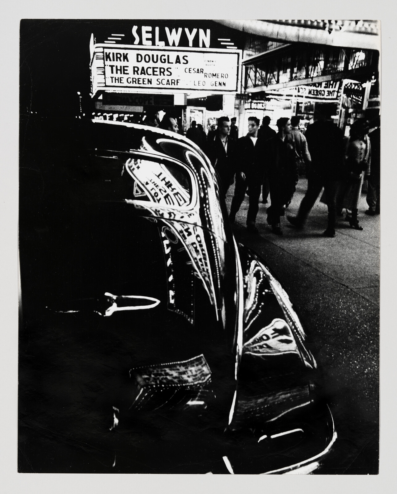 A shiny car hood reflects the Selwyn theater marquee advertising Kirk Douglas films while people walk by.