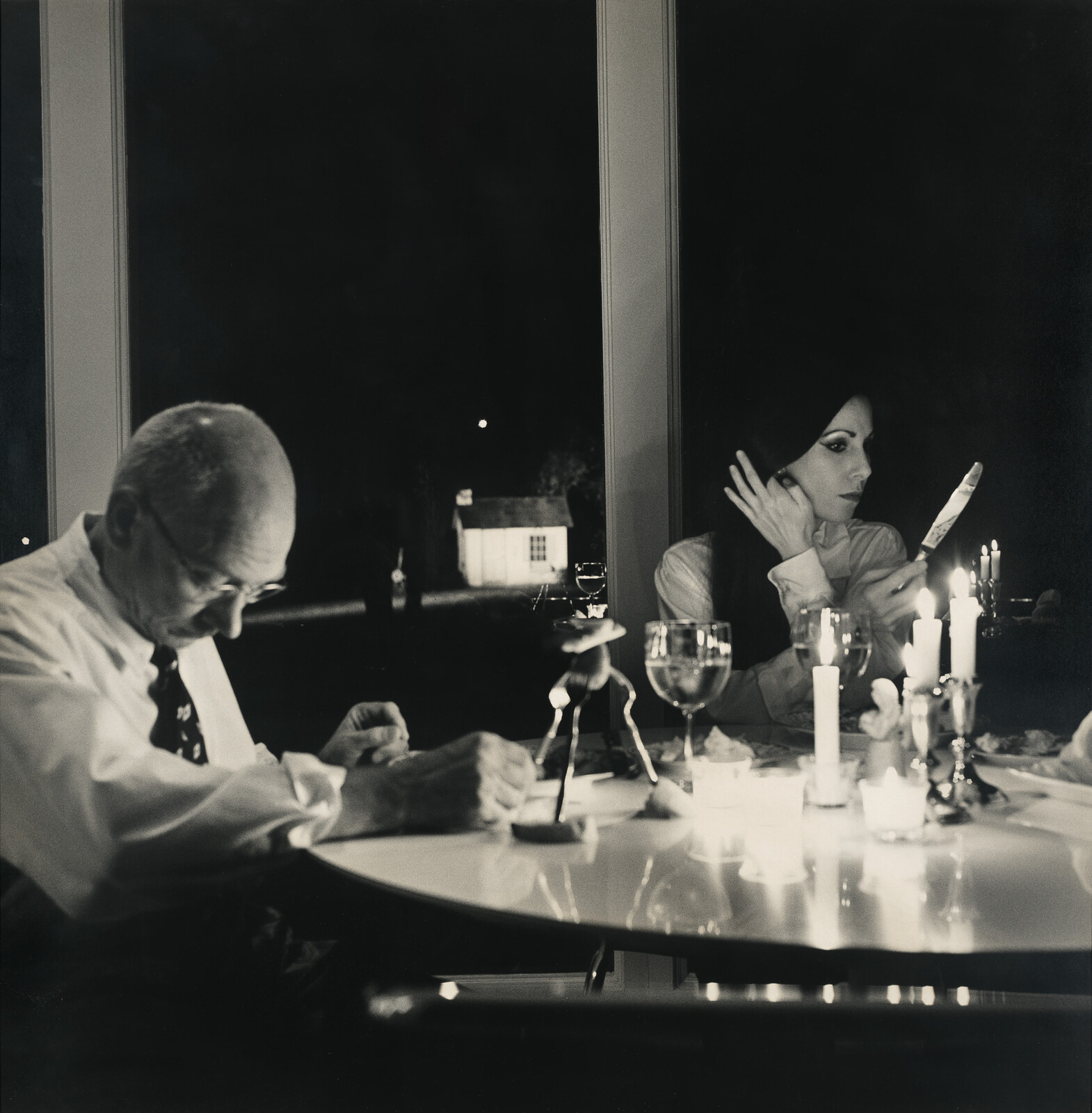 A woman holds a small mirror by candlelight while a man looks down at the table.