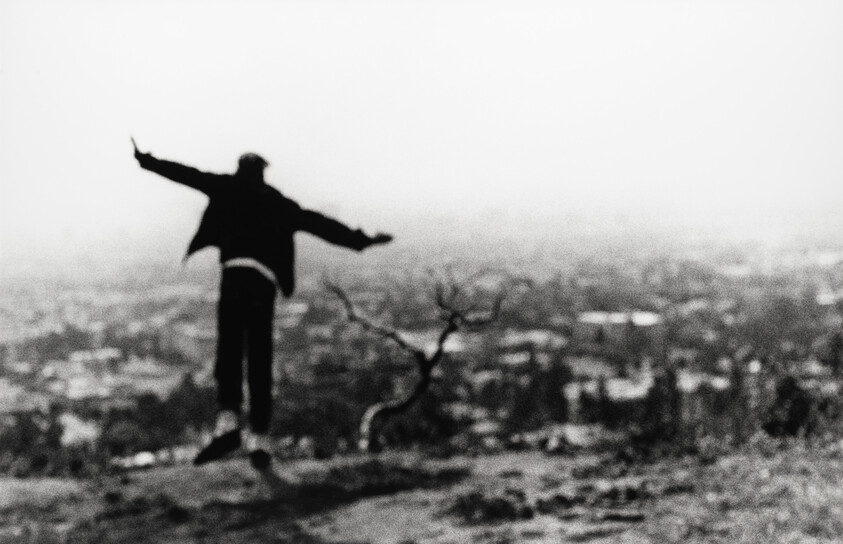 A person balances on one foot with arms outstretched on a hill overlooking a blurred city.
