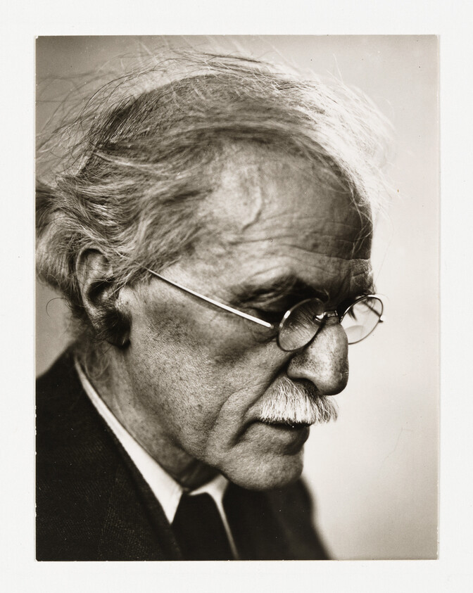 Elderly man with round glasses and white mustache looks down thoughtfully in close-up profile.