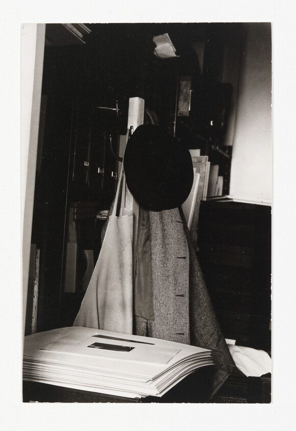 A coat and hat hang on a rack next to a stack of paper prints.