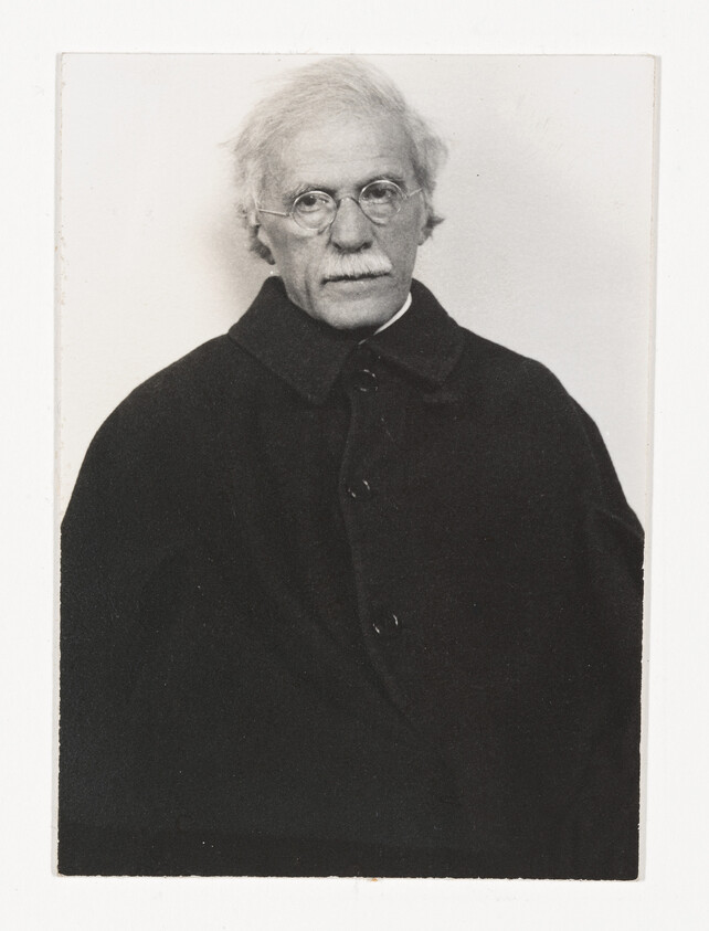 Elderly man with round glasses and white mustache wearing a dark buttoned coat, looking forward.