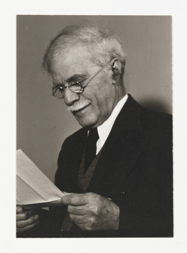 Elderly man with glasses and suit reading a letter and smiling softly.