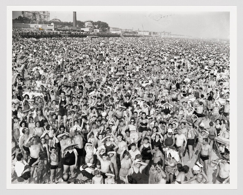 Thousands of beachgoers crowd the shoreline, many shielding their eyes from the sun.