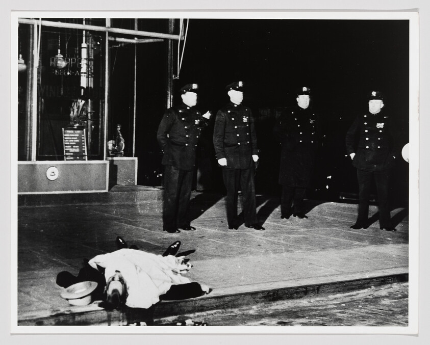 A covered body lies on the sidewalk as four uniformed police officers stand nearby.