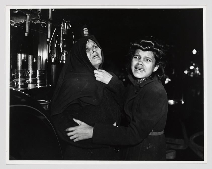 Two distressed women cling to each other, crying and looking toward the camera at night.