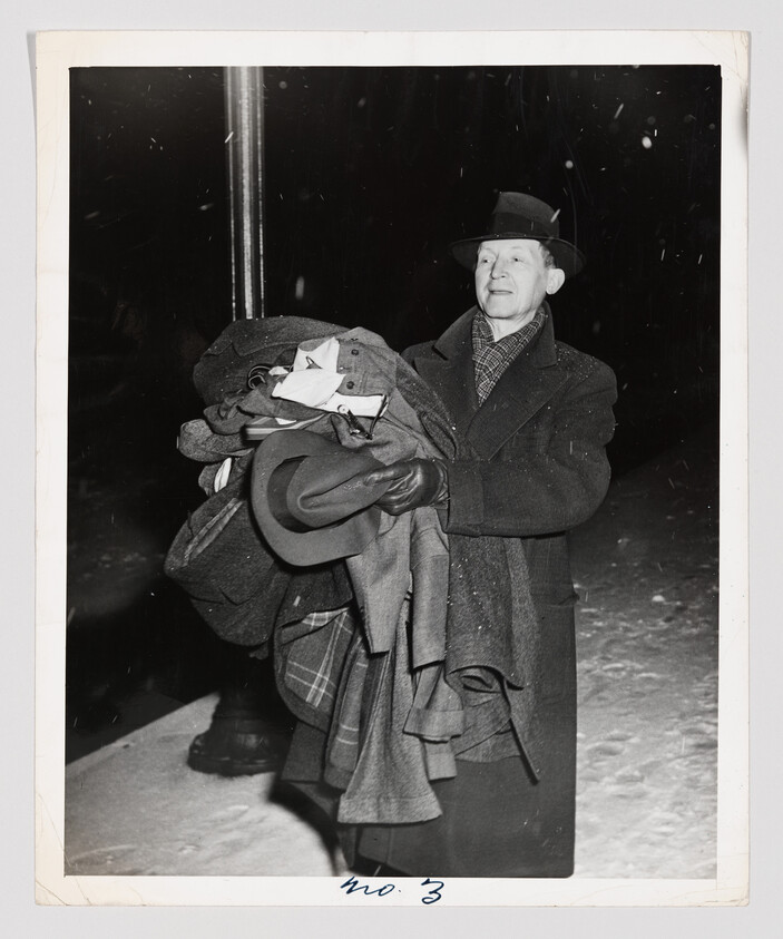 A black and white photograph of a man standing in the snow at night, holding a large bundle of coats or blankets. He is wearing a hat, overcoat, and gloves, and appears to be looking off to the side. Snowflakes are visible in the air around him.