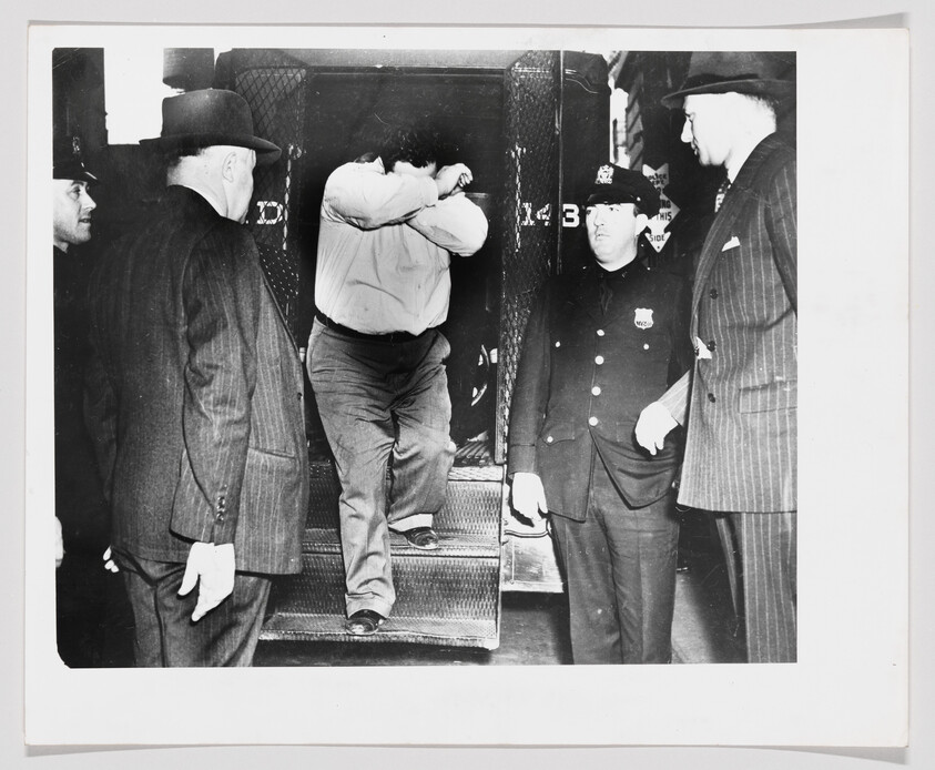 A man covering his face steps down from a police van while officers and detectives watch.