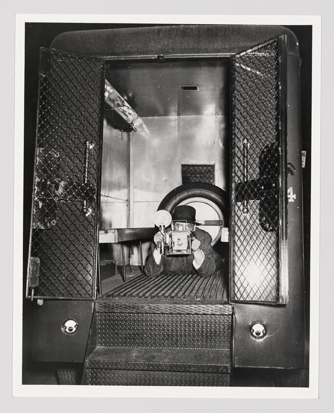 Man lying inside a van aiming an old camera toward the viewer.