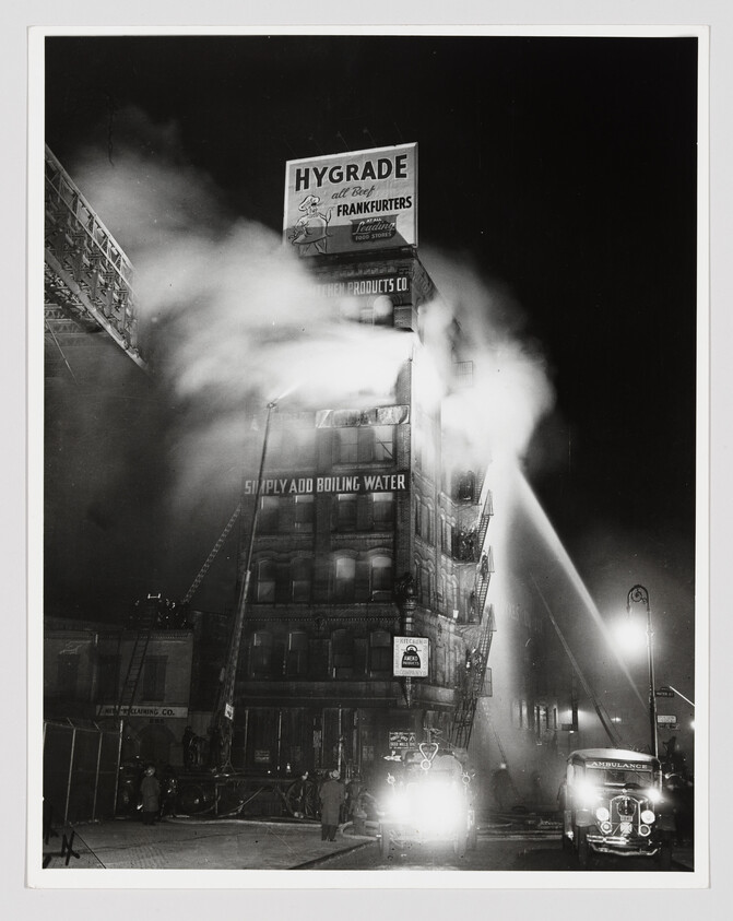 Tall commercial building with "Hygrade" sign surrounded by fire trucks spraying water to fight heavy smoke.