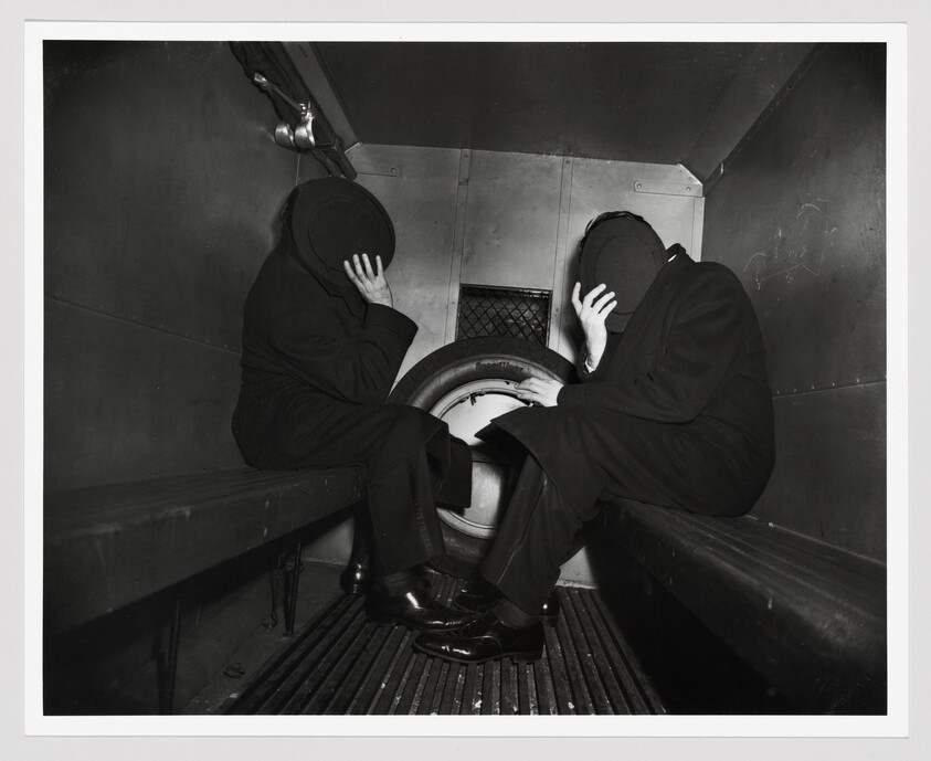 Two individuals in dark clothing and hats sit on a bench inside a vehicle, possibly a train or bus, with their faces obscured by their hands. They appear to be in a state of distress or hiding their identities. The setting is confined and utilitarian, with a metal grate on the floor and a simple bench for seating.