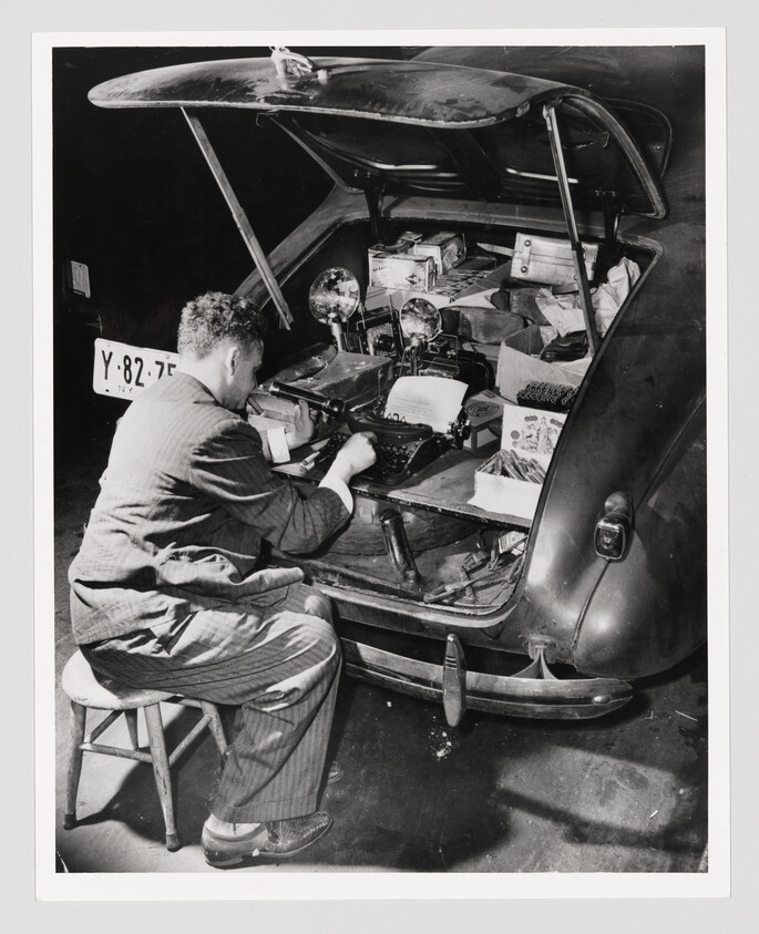 Man seated on a stool typing on a typewriter set up in a car's open trunk.