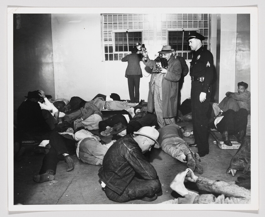 Several men lie on a room floor while a police officer and photographers document them.