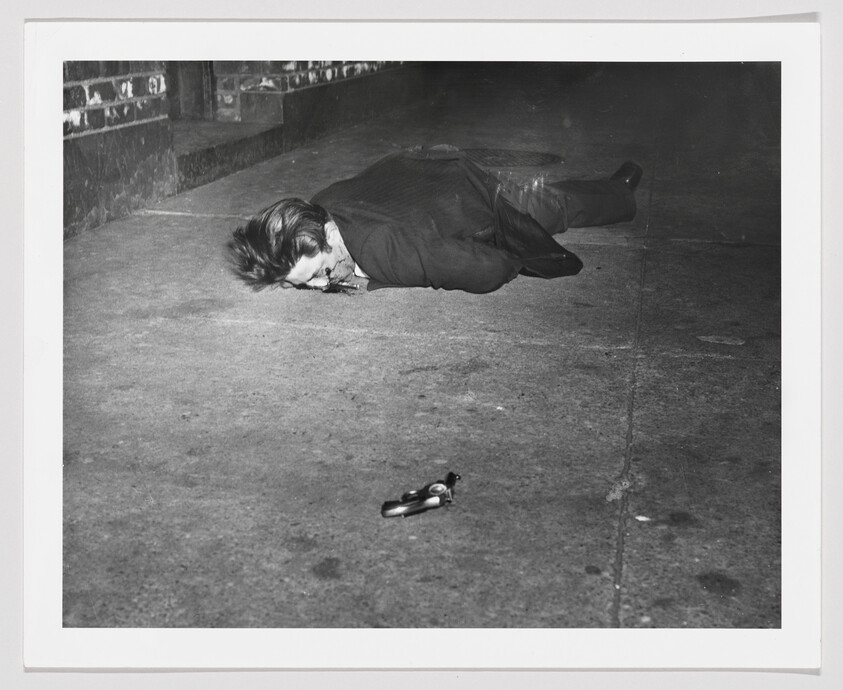 A person lies motionless face down on a sidewalk with a handgun nearby.