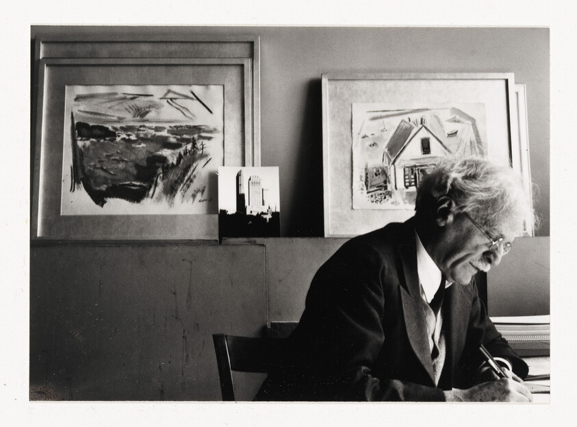 An elderly man in a suit leans over a desk writing with framed paintings behind him.
